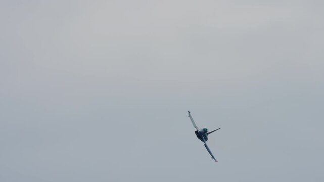 Fighter jet pulling up into a sharp climb, military aircraft banking steeply against an overcast grey sky, combat jet showing its underside during an air display, filmed in slow motion