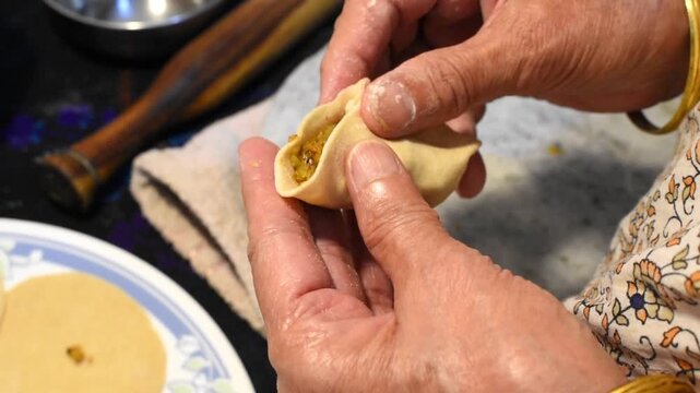 elderly Indian senior lady hands stuffing cabbage filling into whole wheat dough and shaping healthy homemade momo or dumpling in a home kitchen.
