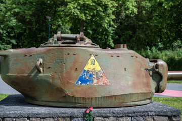 WWII American Sherman tank turret monument with 10th Armored Division insignia in Bastogne, Belgium. Historic military memorial on a stone plinth surrounded by green trees on a sunny day. © Rudolf