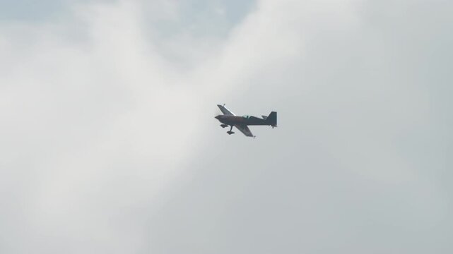 Aerobatic plane cruising in level flight, single engine sport aircraft passing bright hazy clouds, propeller plane with fixed landing gear at an air show, filmed in slow motion