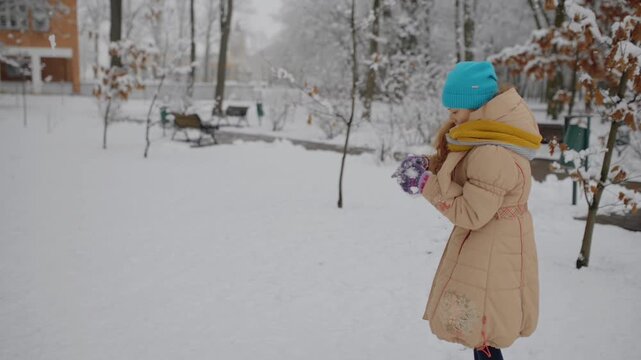 little beautiful girl in a beige jacket sculpts snowballs and throws them in the winter city park