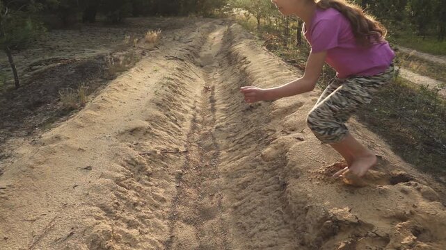 Girl in camouflage pants and pink T-shirt jumps through sandy moat. Slow motion. Evening time, sunset.