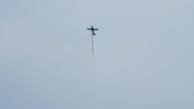 Aerobatic plane powering into a vertical climb, single engine sport aircraft trailing white smoke against a pale blue sky, stunt plane at an air show, filmed in slow motion