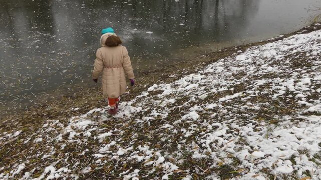 Small beautiful girl in yellow jacket goes to frozen pond in old city park.