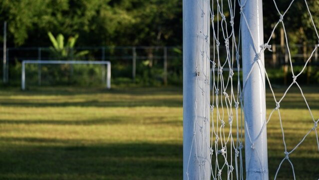 A selective-focus photo featuring an empty sports net, with the faint outline of practice goalposts visible in the distance against a rural backdrop.