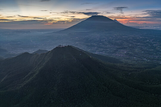 Aerial view of Gunung Telomoyo at sunrise with lush forested slopes, a mountain peak in the distance, and soft morning light in Ngablak, Central Java, Indonesia.