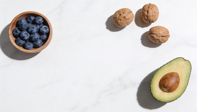 Top view of fresh blueberries in a wooden bowl, walnuts, and half an avocado on a white marble background.