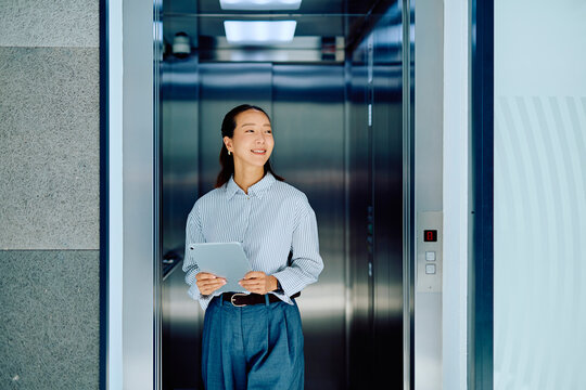 Young Asian businesswoman smiling, holding a digital tablet, and stepping out of a modern elevator in an office building