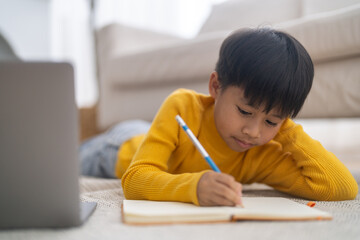 Naklejka premium Asian little boy lying on floor doing homework with laptop online learning, child study, e-learning, homeschooling, smart digital classroom technology and focused childhood educational development