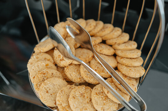 Golden cookies fill wire basket beside serving tongs, shown closeup on dark buffet table for catering use