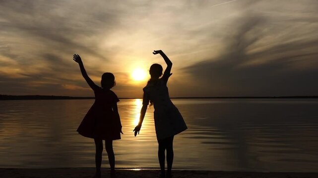 Two little sisters wave their hands in front of the setting sun and a large lake. Beautiful sunset.