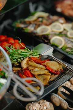 Colorful roasted vegetables fill buffet trays, with tongs beside peppers, potatoes, mushrooms and garnish