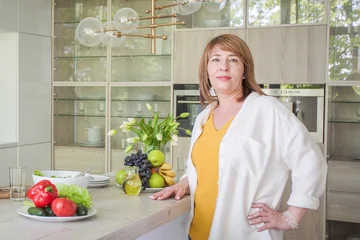 Woman stands in modern kitchen with fresh vegetables and fruits on table during sunny day © artmim