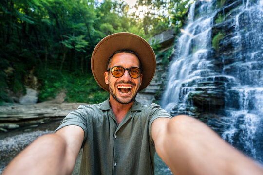 Handsome tourist taking selfie picture in front of waterfall - Happy hiker exploring forest - Traveling life style concept with happy man wearing hat and sunglasses enjoying freedom in the nature