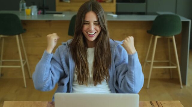 Overjoyed young woman working on her laptop receiving great news, celebrating success with a winning gesture, feeling happy and excited while sitting at a desk in her modern home office
