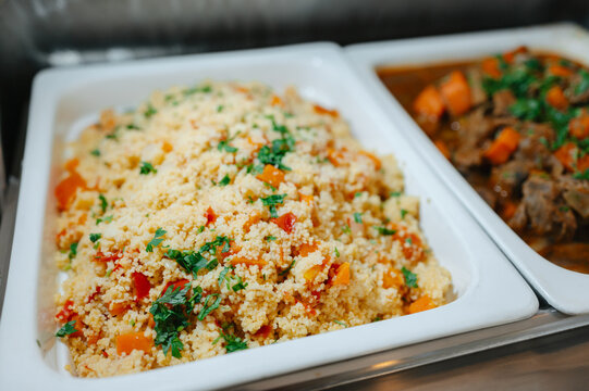 Colorful couscous salad with peppers and herbs, in white buffet trays beside warm cooked vegetables salad