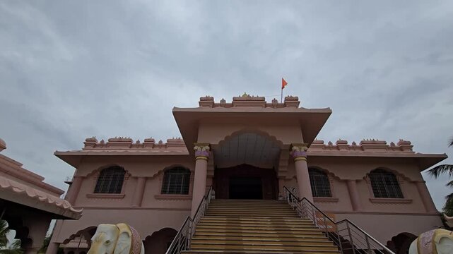 A wide-angle view captures the pink Shri Swami Samarth Seva Kendra temple in Ganagapur, Karnataka. Grand stairs lead up to the entrance, while a vibrant saffron bhagva flag billows from the rooftop.