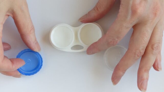 Woman's hands unscrew the lid of a contact lens case filled with contact lens solution. Eye care and hygienic lens storage.