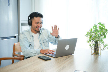 Fototapeta premium Asian man with headphones in kitchen making hand gesture during video call, expressive work communication.