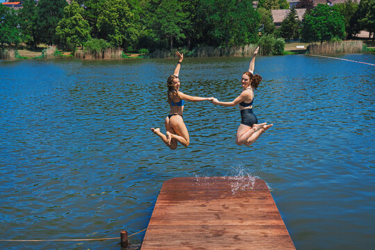 A shot of two attractive young women in bikinis at a small lake. Jumping into the water from a wooden jetty.