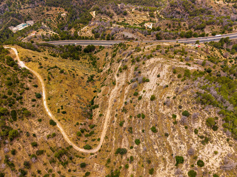 Aerial view of a winding dirt road cutting through a hilly landscape with sparse vegetation, pine trees, and a highway running along the ridge in Calp, Valencian Community, Spain.