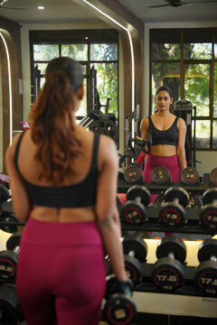 Maharashtra, Sion, India- 22nd March 2026- Young Indian girl  in her active wear doing workout with dumbbells in gym, her reflection is seen in the mirror.