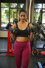 Fototapeta premium Maharashtra, Sion, India- 22nd March 2026- Young Indian girl in her active wear doing workout with two dumbbells in gym.