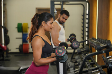 Fototapeta premium Maharashtra, Sion, India- 22nd March 2026- Young Indian girl and guy doing workout with dumbbells, looking into the mirror in gym.