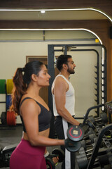 Fototapeta premium Maharashtra, Sion, India- 22nd March 2026- Young Indian girl and guy doing workout with dumbbells, looking into the mirror in gym.
