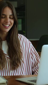 Beautiful young professional woman sitting at her desk, engaging in a pleasant video conference on her laptop, smiling and talking while working from her modern home office
