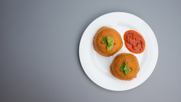 Two vada pav portions with spicy condiments on white plate featuring a matte gray background