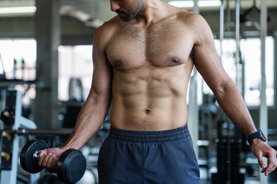 Muscular shirtless man lifting dumbbell doing bicep curl exercise inside fitness gym. Athletic male bodybuilder showing abs sixpack torso during intense workout training session body care concept