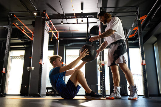 Personal trainer handing medicine ball to young athlete during core workout. Concept of partner training, abs exercise, fitness coaching and gym routine.