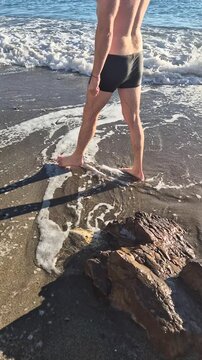 Man beach walking, shirtless male in swimwear enjoying the sunny ocean waves on a sandy shore during summer. Vertical video
