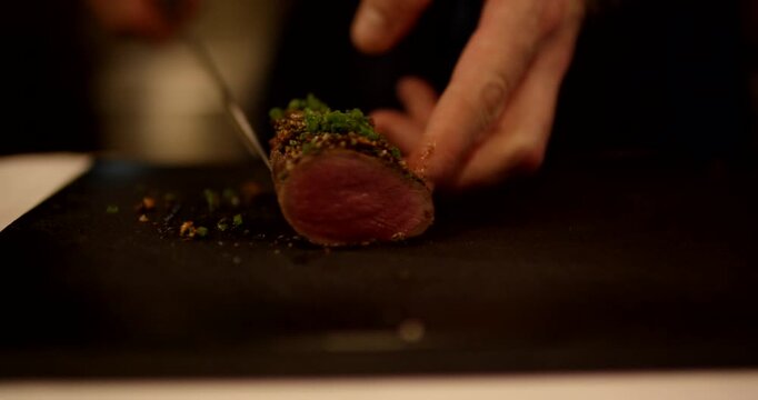 Close up view of a chef slicing a tender piece of medium rare meat topped with green herbs on a dark board