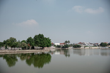 Fototapeta premium Scenic view of a calm urban lake surrounded by lush green trees and residential houses under a clear hazy sky.