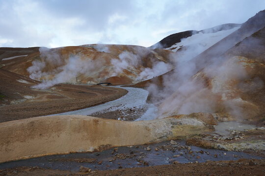 View of the geothermal landscape of Kerlingarfjoll with steaming vents, colorful rhyolite mountains, and a winding stream under a cloudy sky in Hella, Rangarthing ytra, Iceland.