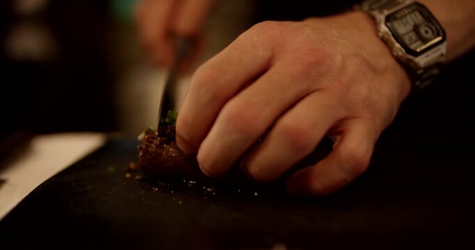 Close up view of hands using a knife to slice a roasted meat piece covered with green herbs on a dark surface