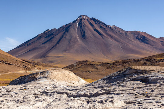 View of the volcanic landscape at Piedras Rojas with rugged rock formations, arid terrain, and a prominent mountain peak under a clear blue sky in San Pedro de Atacama, Antofagasta, Chile.