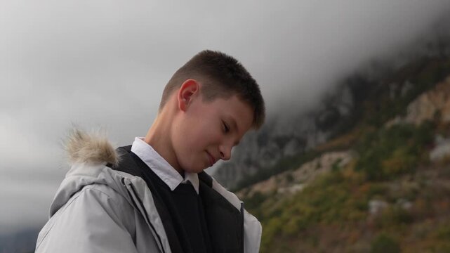 Teenager student hiking against foggy mountains wearing a gray winter coat and feeling sad or pensive outdoors.
