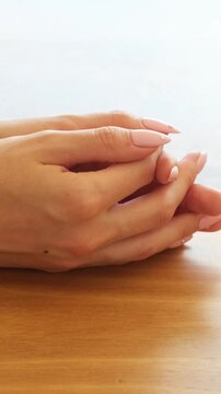 Woman's hands with a fresh nude manicure are resting on a wooden table, with her fingers interlaced in a relaxed and thoughtful gesture showing beautiful, well-cared for skin and nails