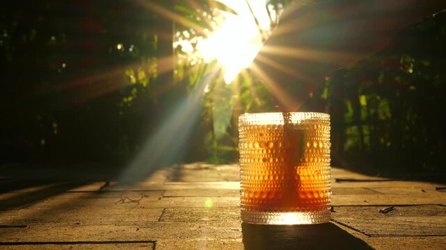 Golden sunbeams stream through leafy trees. Textured glass holds amber liquid and a bright orange garnish. Hand gently places the garnish into the glass