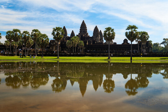 View of the ancient Angkor Wat temple complex with its towers reflected in the moat, framed by palm trees under a blue sky in Krong Siem Reap, Siem Reap Province, Cambodia.