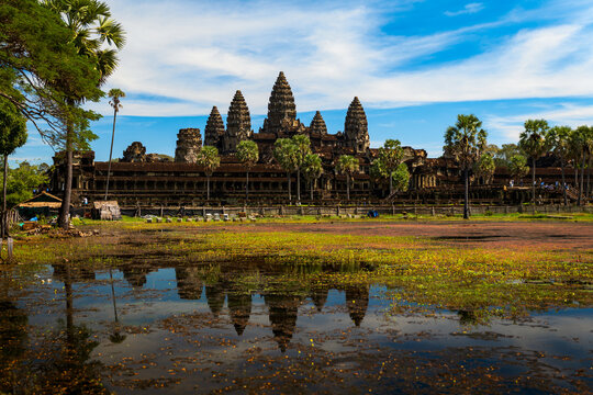 View of the ancient Angkor Wat temple complex reflected in the foreground pond, surrounded by tall palm trees under a bright blue sky in Krong Siem Reap, Siem Reap Province, Cambodia.