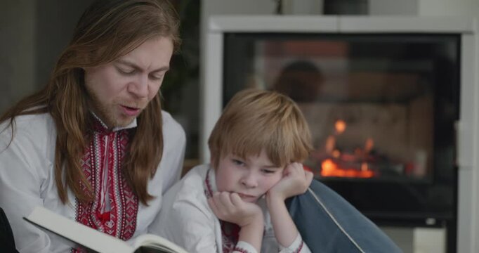 Man Reads Book with Kid Boy Together on Floor Near Fireplace. Happy Family in Ukrainian Traditional Attire. 4K 10-bit