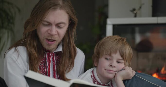 Man Reads Book with Kid Boy Together on Floor Near Fireplace. Happy Family in Ukrainian Traditional Attire. 4K 10-bit