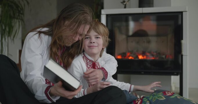 Man Reads Book with Kid Boy Together on Floor Near Fireplace. Happy Family in Ukrainian Traditional Attire. 4K 10-bit