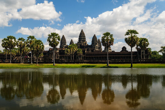 View of Angkor Wat temple complex reflected in the surrounding moat with palm trees under a blue sky with clouds in Krong Siem Reap, Siem Reap Province, Cambodia.