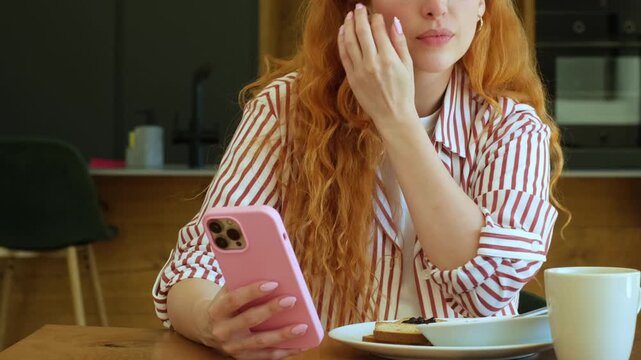 Beautiful redhead woman with long curly hair browsing her smartphone and drinking coffee during breakfast in the kitchen, checking social media and enjoying a relaxed morning at home