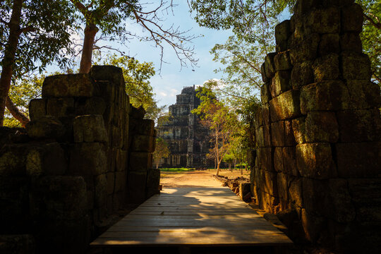 View of the ancient stone temple ruins framed by lush trees and a wooden walkway in Krong Siem Reap, Siem Reap Province, Cambodia.